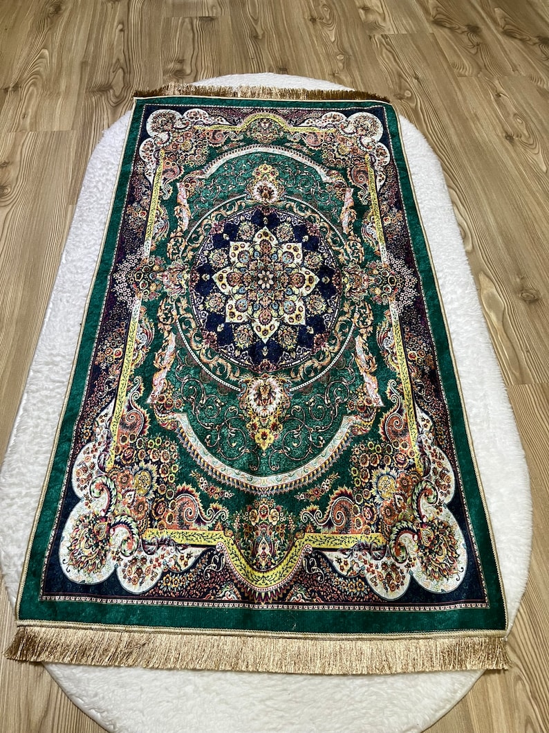 Peut inclure: Un tapis de pri&egrave;re vert avec des motifs floraux et g&eacute;om&eacute;triques complexes dans des tons d'or, de jaune, de rouge et de bleu. Le tapis a une bordure de franges dor&eacute;es.