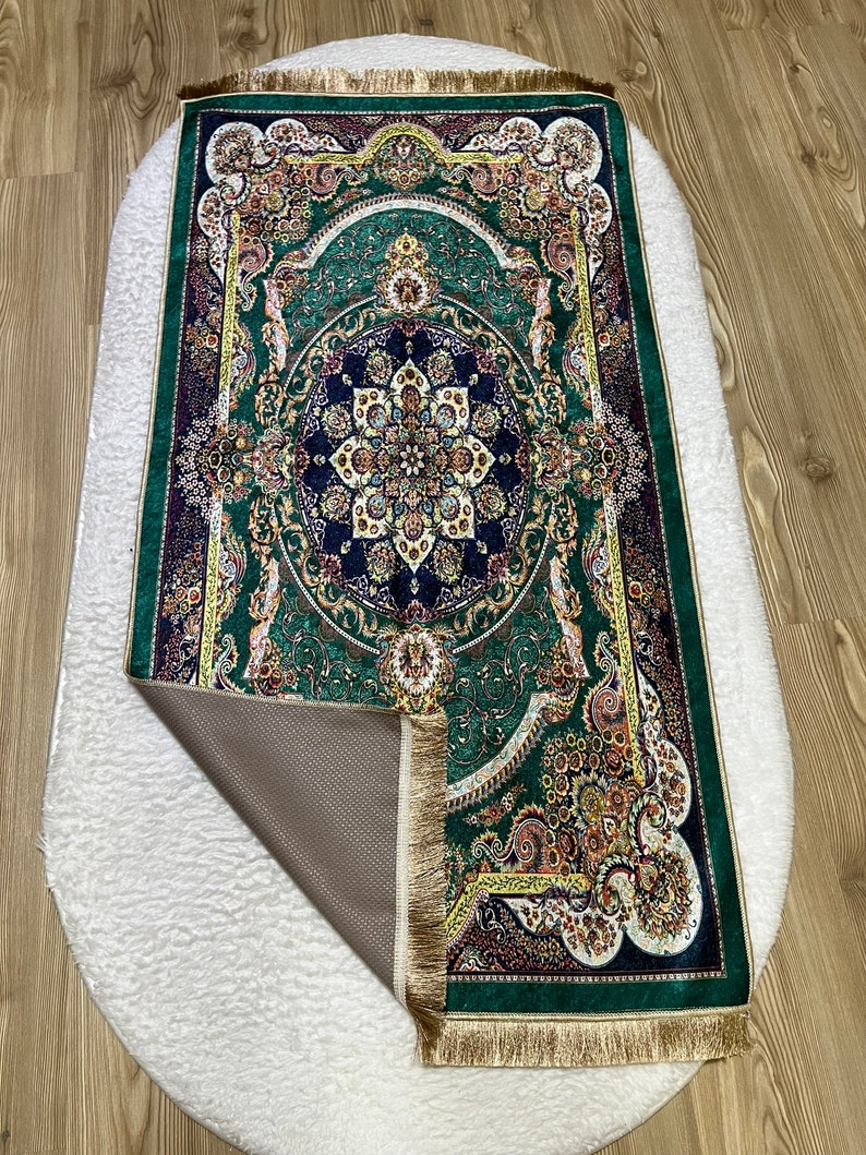 Peut inclure: Un tapis de pri&egrave;re vert avec des motifs floraux et dor&eacute;s complexes. Le tapis a une bordure frang&eacute;e et un dos antid&eacute;rapant.