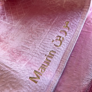 May include: A pink prayer rug with a gold embroidered border and the name "Maauim" embroidered in gold.