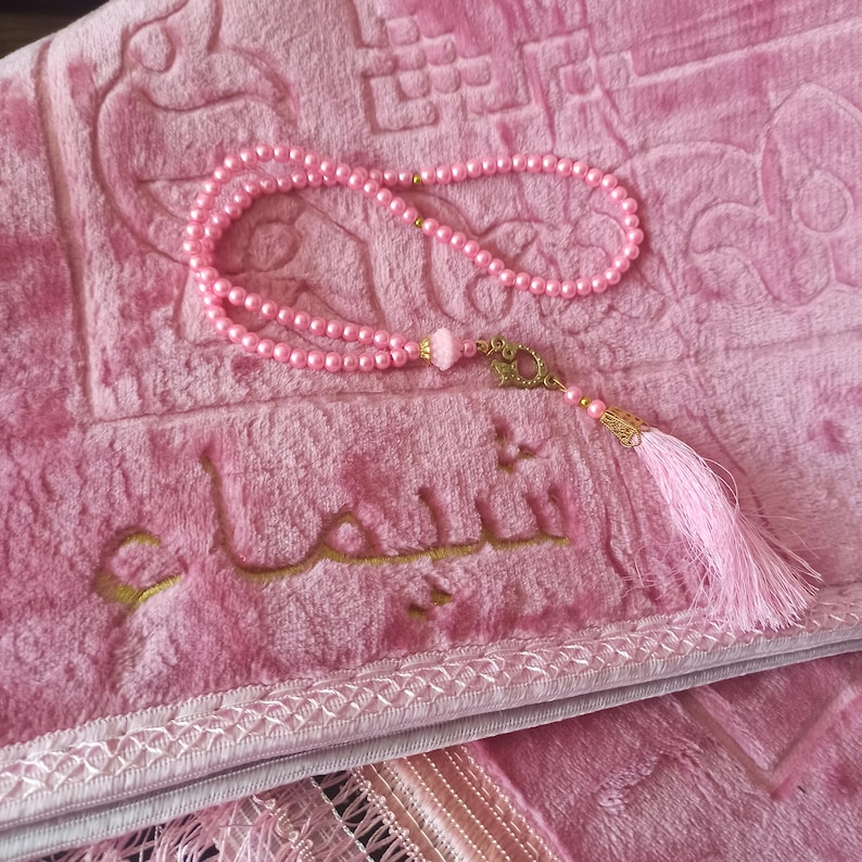 May include: A pink prayer rug with a gold embroidered inscription in Arabic. A pink prayer bead necklace with a gold tassel hangs on the rug.