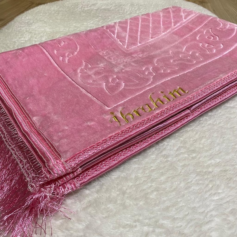 May include: Pink prayer rug with a gold embroidered design and the word "Ibrahim" in gold lettering. The rug has a fringe border.