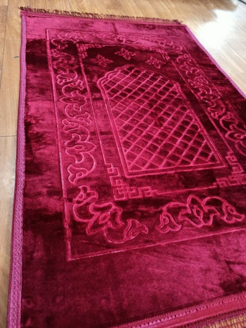 May include: A burgundy prayer rug with intricate embroidered designs and a central prayer space. The rug has a soft, plush texture and a fringed border.