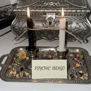 May include: A silver tray holds a black and white candle, connected by twine, with flames lit. The tray also contains dried flowers and a card that reads "PSYCHIC INDIGO". The words "Cord Cutting Ritual" are at the top.