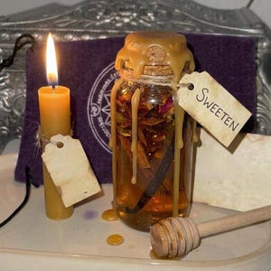 May include: A lit beeswax candle and a glass jar filled with herbs, spices, and a vanilla bean. The jar is labeled "Sweeten" and has a caramel-colored wax seal. A wooden honey dipper rests nearby.