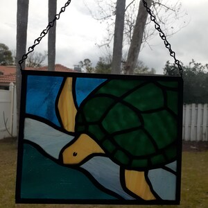 May include: Stained glass panel featuring a turtle swimming in blue and green water. The turtle has a green shell, yellow head and flippers. The panel is framed in black and suspended by a chain.