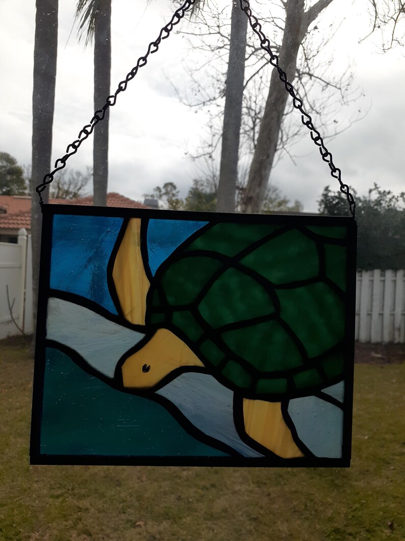 May include: Stained glass panel featuring a turtle swimming in blue and green water. The turtle has a green shell, yellow head and flippers. The panel is framed in black and suspended by a chain.