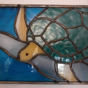 May include: Stained glass panel featuring a sea turtle design. The turtle has a green shell, yellow head, and is set against a blue and white background. The glass is framed with a dark metal border.