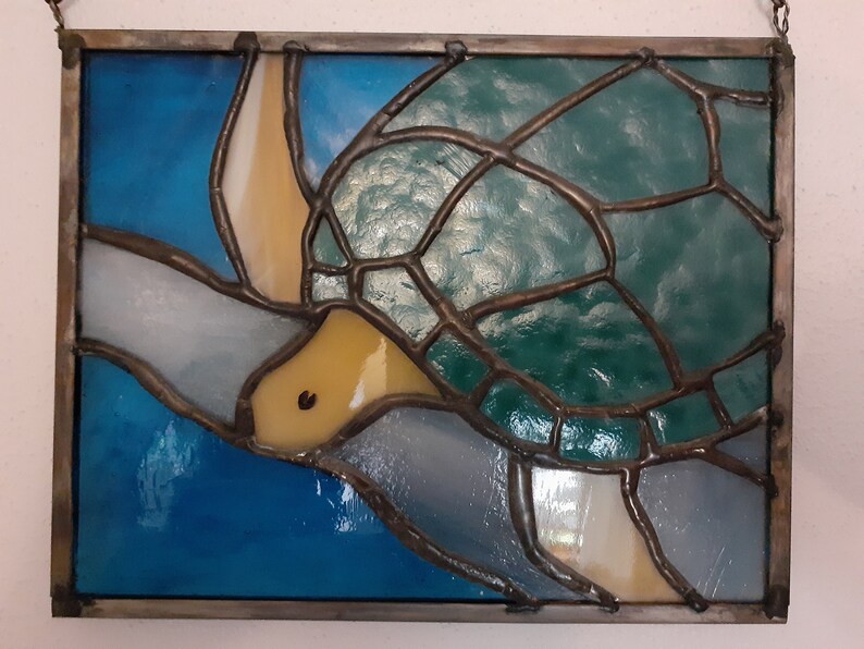 May include: Stained glass panel featuring a sea turtle design. The turtle has a green shell, yellow head, and is set against a blue and white background. The glass is framed with a dark metal border.