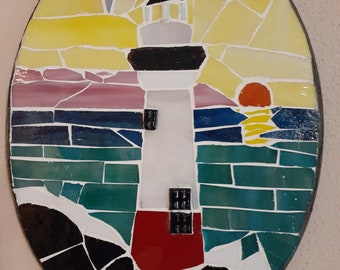 May include: Oval mosaic art piece depicting a lighthouse scene. The lighthouse is white with a black top and red base, set against a sunset with yellow, pink, and blue hues. The mosaic is framed with a dark border.
