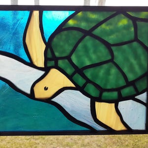 May include: Stained glass panel featuring a sea turtle design. The turtle has a green shell, yellow head and flippers, and is set against a blue and white background. The panel is framed in black.