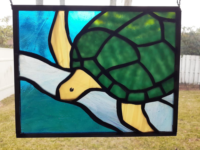 May include: Stained glass panel featuring a sea turtle design. The turtle has a green shell, yellow head and flippers, and is set against a blue and white background. The panel is framed in black.