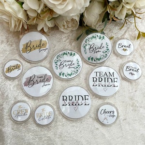 Könnte beinhalten: Sortiment runder, weißer Plastikabzeichen mit grünem Laub, goldenen Akzenten und schwarzem Text. Die Abzeichen zeigen die Wörter "Bride", "Team Bride", "Cheers" und "The Crew".