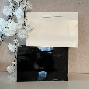 Puede incluir: Dos bolsas de regalo, una negra y una blanca, apiladas una encima de la otra. La bolsa blanca tiene un asa de cuerda blanca.
