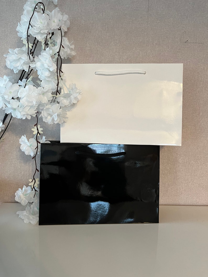Puede incluir: Dos bolsas de regalo, una negra y una blanca, apiladas una encima de la otra. La bolsa blanca tiene un asa de cuerda blanca.