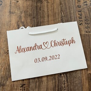 Puede incluir: Una bolsa de regalo blanca con un asa de cuerda blanca. La bolsa tiene una inscripci&oacute;n de color cobre que dice "Alexandra & Christoph 03.09.2022".