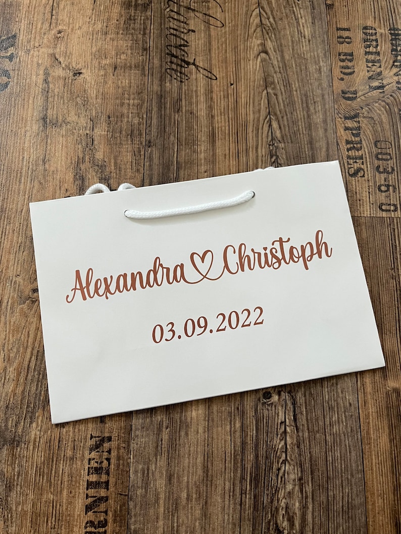 Puede incluir: Una bolsa de regalo blanca con un asa de cuerda blanca. La bolsa tiene una inscripci&oacute;n de color cobre que dice "Alexandra & Christoph 03.09.2022".