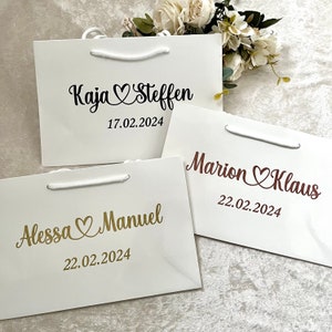Puede incluir: Tres bolsas de regalo blancas con asas. Las bolsas est&aacute;n decoradas con letras doradas y rojas y corazones. El texto en las bolsas dice "Kaja & Steffen 17.02.2024", "Marion & Klaus 22.02.2024" y "Alessa & Manuel 22.02.2024".