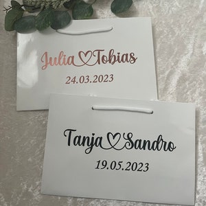 Puede incluir: Dos bolsas de regalo blancas con asas. La bolsa de arriba tiene el texto "Julia Tobias" con un s&iacute;mbolo de coraz&oacute;n y la fecha "24.03.2023". La bolsa de abajo tiene el texto "Tanja Sandro" con un s&iacute;mbolo de coraz&oacute;n y la fecha "19.05.2023".