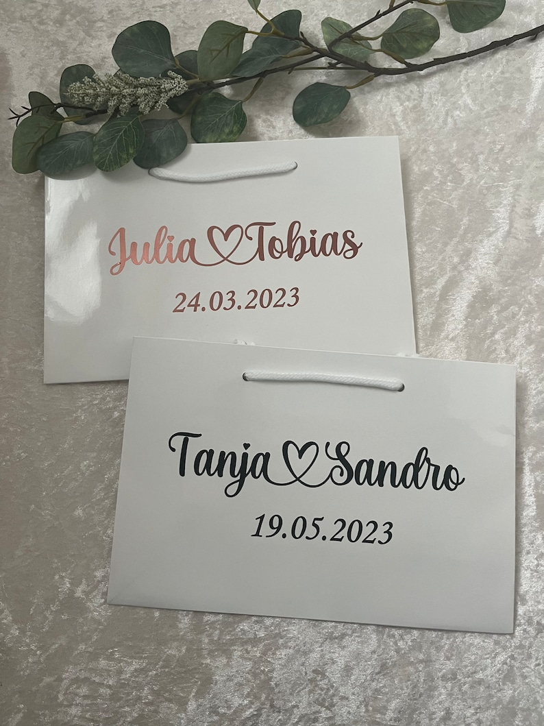 Puede incluir: Dos bolsas de regalo blancas con asas. La bolsa de arriba tiene el texto "Julia Tobias" con un s&iacute;mbolo de coraz&oacute;n y la fecha "24.03.2023". La bolsa de abajo tiene el texto "Tanja Sandro" con un s&iacute;mbolo de coraz&oacute;n y la fecha "19.05.2023".