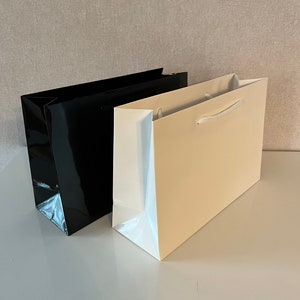 Puede incluir: Dos bolsas de regalo, una negra y otra beige, con asas blancas. Las bolsas est&aacute;n sobre una superficie blanca.