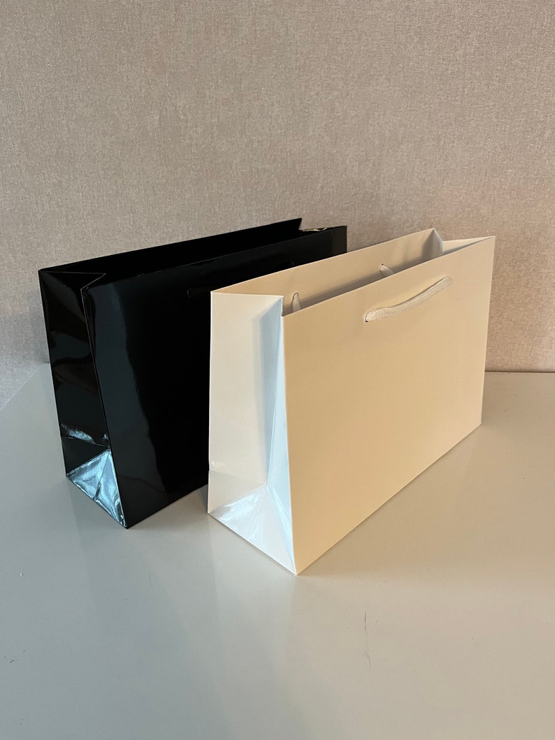 Puede incluir: Dos bolsas de regalo, una negra y otra beige, con asas blancas. Las bolsas est&aacute;n sobre una superficie blanca.