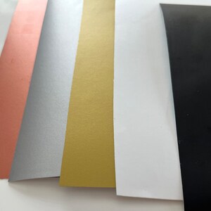 Puede incluir: Cinco hojas de papel en diferentes colores: oro rosa, plata, oro, blanco y negro.