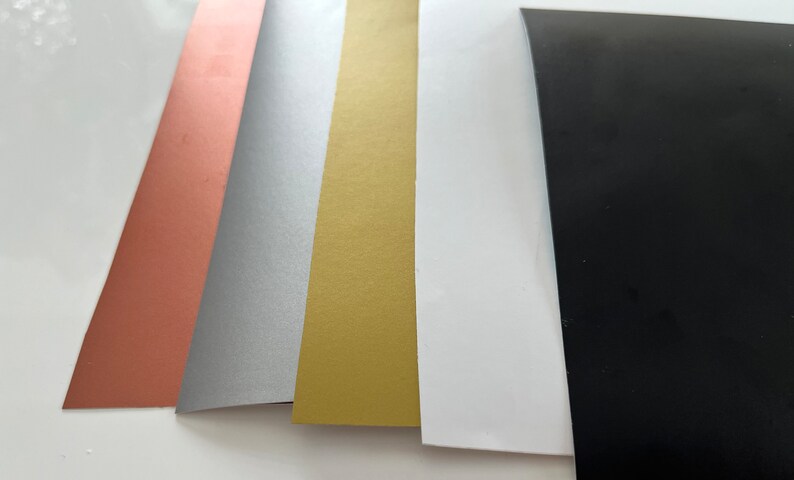 Puede incluir: Cinco hojas de papel en diferentes colores: oro rosa, plata, oro, blanco y negro.
