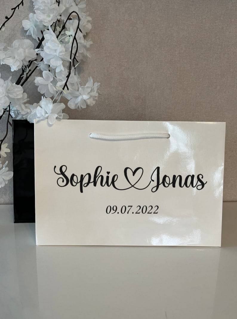 Puede incluir: Una bolsa de regalo blanca con asas negras y una inscripci&oacute;n negra que dice "Sophie &hearts; Jonas 09.07.2022".