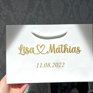 Puede incluir: Bolsa de regalo blanca con una inscripci&oacute;n dorada que dice "Lisa &hearts; Mathias 11.08.2022".