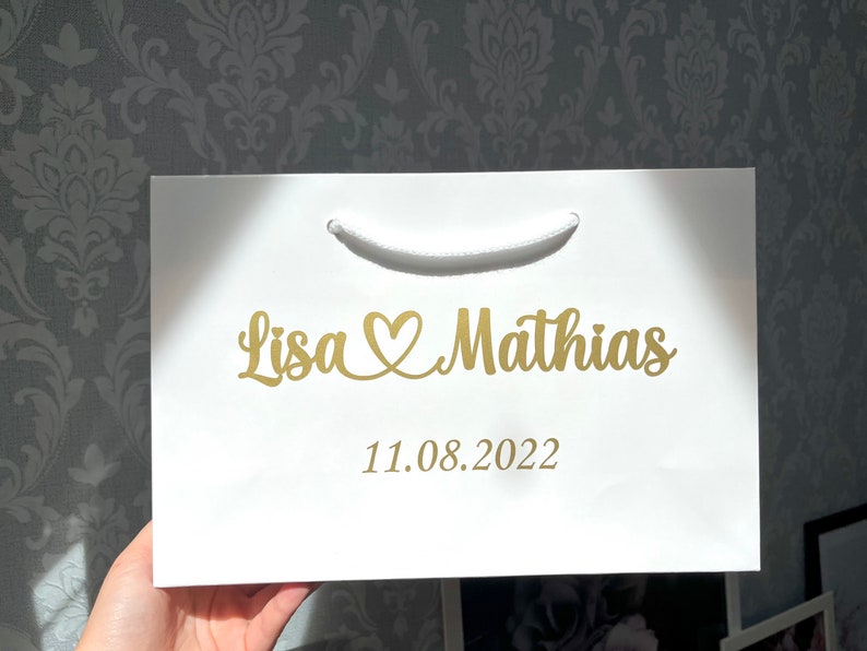 Puede incluir: Bolsa de regalo blanca con una inscripci&oacute;n dorada que dice "Lisa &hearts; Mathias 11.08.2022".