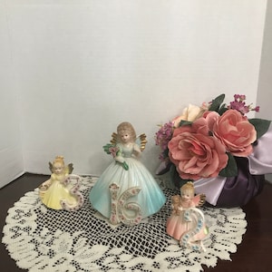 May include: Three porcelain angel figurines holding numbers 3, 16, and 2, with floral details. The angel figurines wear dresses in yellow, blue, and pink. A bouquet of pink and purple flowers is in the background.