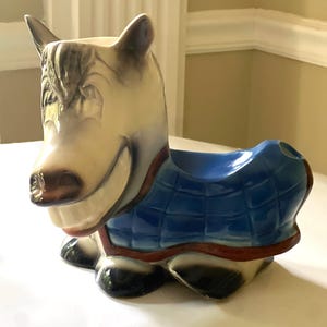 Peut inclure: Pot de fleurs en céramique en forme de cheval souriant. Le cheval a un corps crème, une crinière et des oreilles grises, et une couverture matelassée bleue. Le pot a une bordure rouge et des sabots noirs.