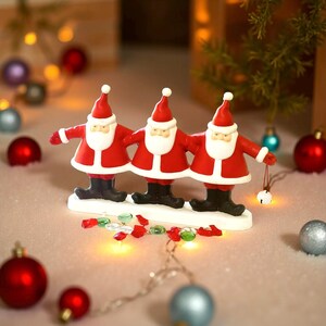 May include: A festive Christmas decoration featuring three Santa Claus figurines in red suits, white beards, and black boots. They stand on a white base, surrounded by ornaments and lights, creating a cheerful holiday scene.