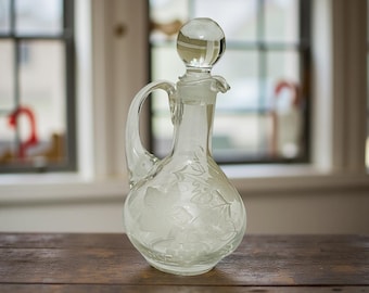 Op de afbeelding: Een heldere glazen karaf met een ronde basis, een handvat en een stop. De karaf heeft geëtste bloemmotieven en staat op een houten oppervlak. De achtergrond bevat een raam met natuurlijk licht.