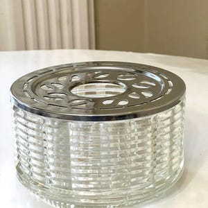 May include: A clear glass tea warmer with a silver metal top. The top has a decorative design with cutouts. The warmer has a ribbed design.