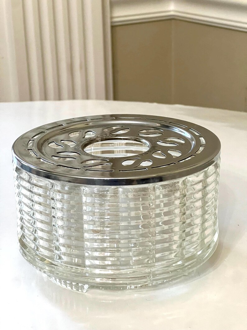 May include: A clear glass tea warmer with a silver metal top. The top has a decorative design with cutouts. The warmer has a ribbed design.