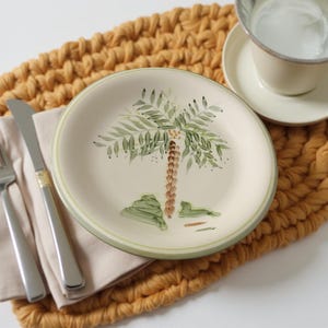 May include: A cream-colored plate with a hand-painted palm tree design. The plate has a green border and is set on a woven yellow placemat. A fork, knife, and teacup are also visible.
