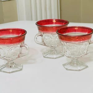 Puede incluir: Tres tazas de té de vidrio transparente con bordes y asas rojas. Las tazas tienen un diseño decorativo y se asientan sobre bases hexagonales. Las tazas de té están dispuestas sobre una superficie blanca.