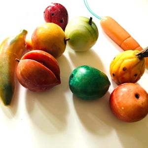 Könnte beinhalten: Eine Sammlung farbenfroher, dekorativer Steinfrüchte und -gemüse. Enthält eine Banane, einen Apfel, eine Karotte und andere Früchte in Rot-, Gelb-, Orange- und Grüntönen. Die Objekte sind auf einer weißen Oberfläche angeordnet.