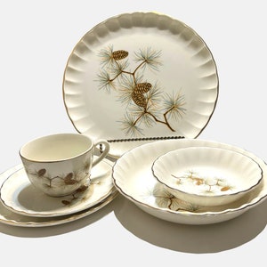 May include: A set of white ceramic dinnerware with a pine cone design. The set includes a dinner plate, a salad plate, a bowl, a cup, and a saucer.