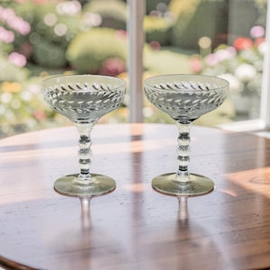 May include: Two vintage champagne coupe glasses in a grey-green hue. The bowls have a textured, diamond pattern, and the stems have a series of small, round beads. The glasses sit on a round base. The glasses are on a wooden table.