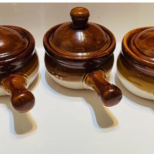 May include: A set of three round ceramic crocks with lids and single handles. Each crock features a two-tone design with a dark brown glazed upper section and a lighter cream-colored base. The lids have a matching dark brown glossy finish.