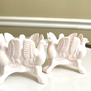 May include: Two white ceramic candle holders shaped like doves with outstretched wings. Each holder has three legs and a circular top.