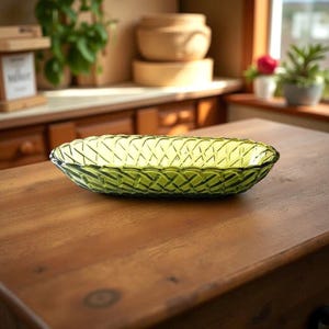 May include: A green glass serving dish with a woven pattern. The oval-shaped dish is made of transparent green glass and sits on a wooden table. The background includes a wooden cabinet and plants.