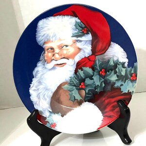 May include: A blue ceramic plate with a festive image of Santa Claus holding a sprig of holly. Santa is wearing a red hat and a white beard.