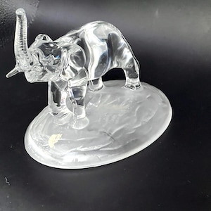 May include: A clear glass elephant figurine with its trunk raised, standing on a frosted oval base. The elephant is detailed with visible wrinkles and folds, showcasing a realistic design. The figurine is set against a dark background.