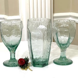 Op de afbeelding: Drie heldere glazen drinkglazen met een getextureerd, mat oppervlak. De glazen zijn groen van kleur en hebben verschillende vormen, waaronder een kelk, een hoog glas en een lager glas.