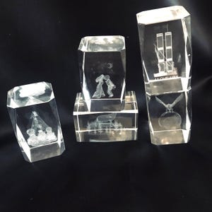 May include: Four clear glass blocks with 3D laser-etched designs. The designs include a couple kissing, a cityscape, a bird, and a globe. The blocks are stacked on top of each other.