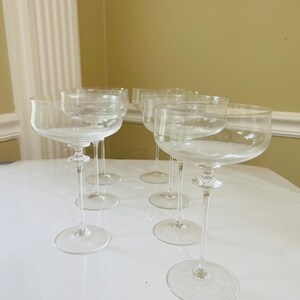 May include: Set of six clear glass champagne coupes with a wide bowl and a long, thin stem. The glasses are arranged in a semi-circle on a white surface.