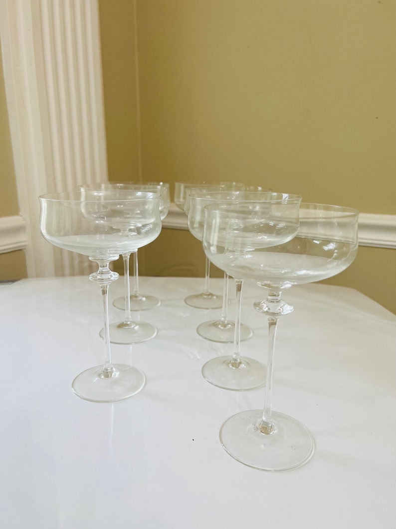 May include: Set of six clear glass champagne coupes with a wide bowl and a long, thin stem. The glasses are arranged in a semi-circle on a white surface.
